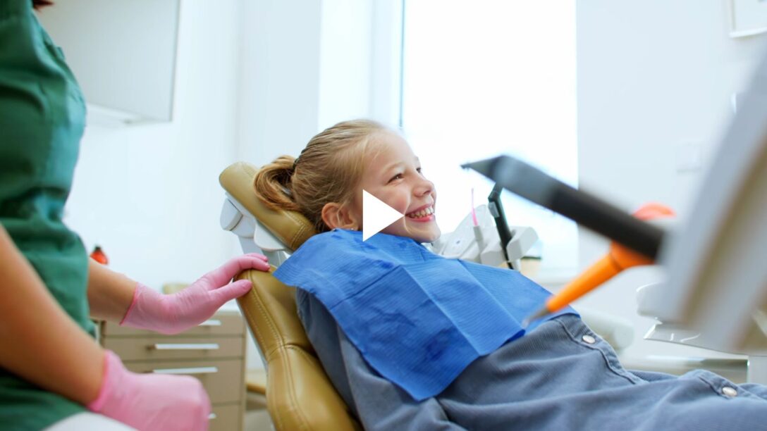 Ein kleines, blondes Mädchen sitzt lachend auf einem ockerfarbenen Behandlungsstuhl beim Zahnarzt.